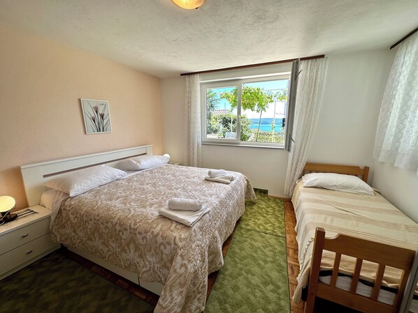A hotel room with a large bed, a smaller bed, a nightstand, a lamp, a window with a view of the sea, and a painting on the wall. - Apartman Mona 2 (Dramalj)