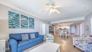 A spacious living room with a blue sofa, a white coffee table, and a ceiling fan. There is a kitchen area in the background.