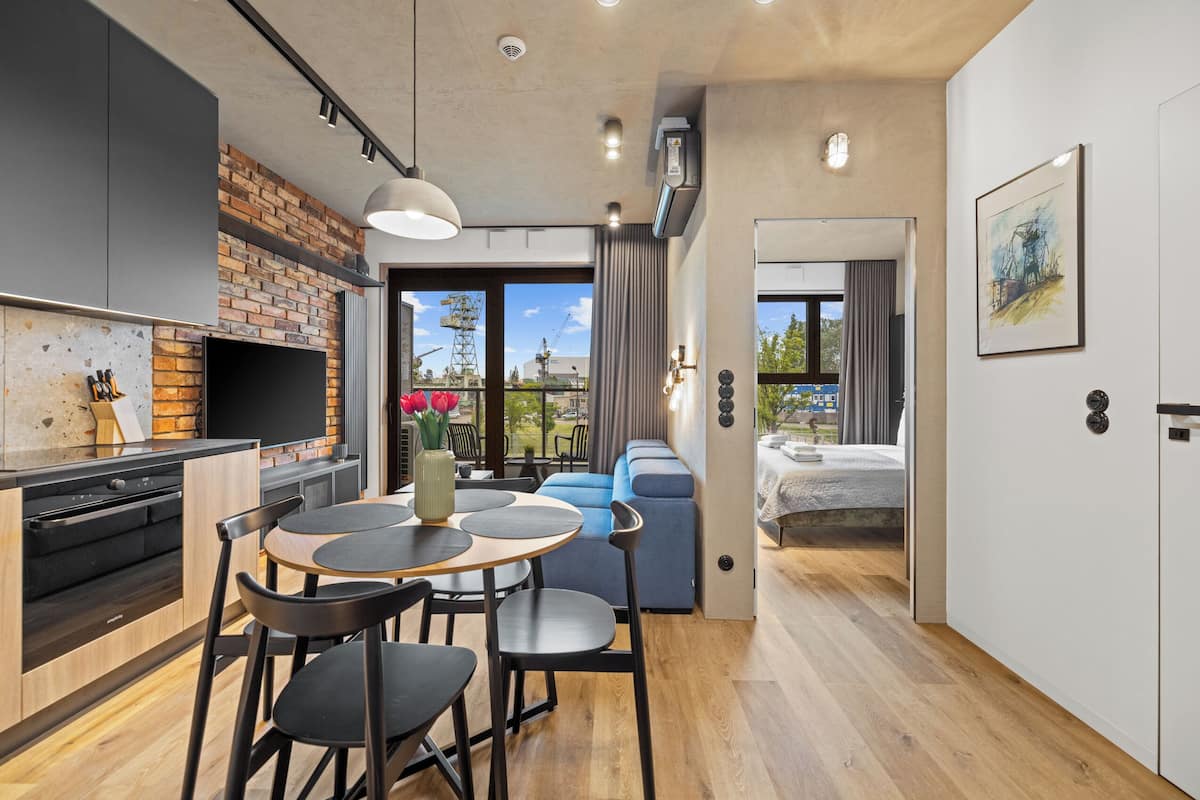 A modern kitchen with a brick backsplash, a dining area with a round table and chairs, and a living area with a sofa and a view of the outdoors.