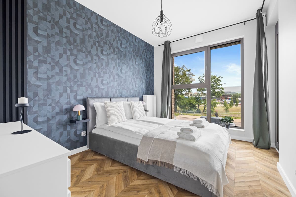 A modern bedroom with a large bed, a bedside table, a lamp, and a view of the outdoors through a large window.