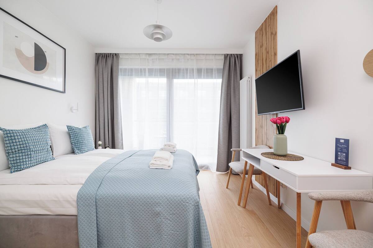 A modern hotel room with a large bed, a desk, a TV, and a window with curtains.