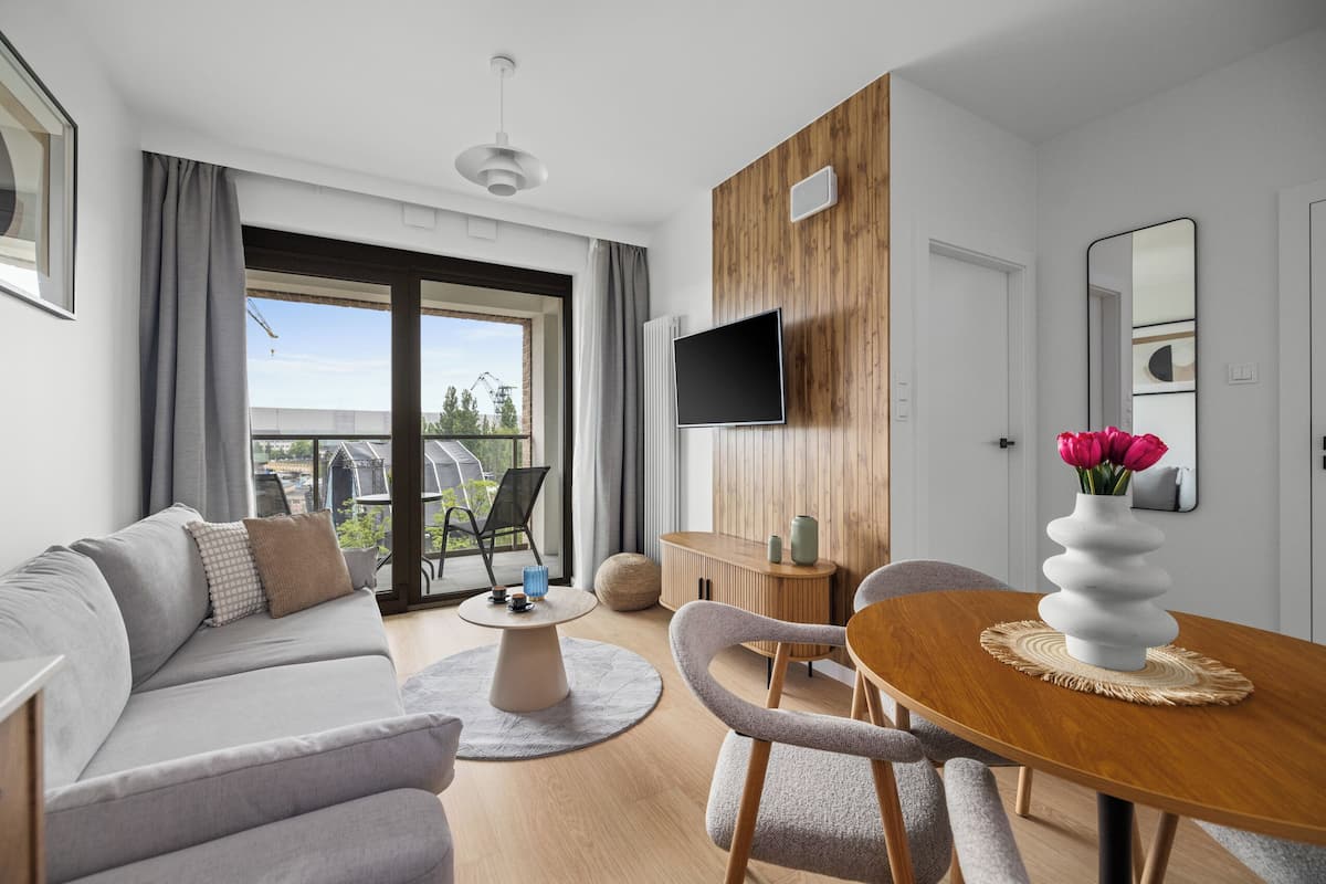 A modern living room with a grey sofa, a wooden dining table, and a TV mounted on the wall.