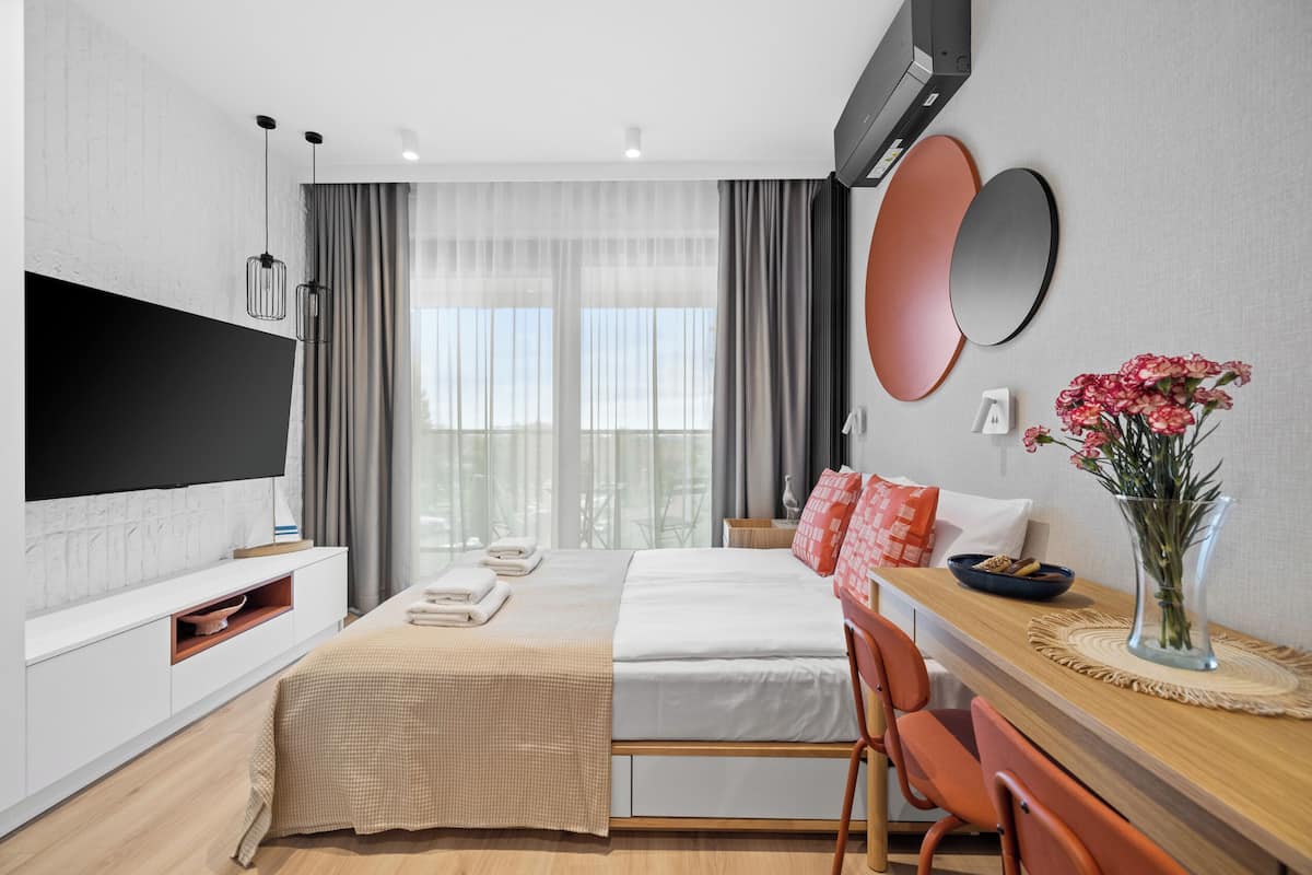 A modern hotel room with a flat-screen TV, a bed with white linens, a wooden dining table, and a vase with flowers.