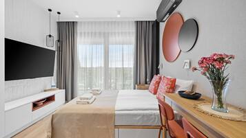 A modern hotel room with a flat-screen TV, a bed with white linens, a wooden dining table, and a vase with flowers.
