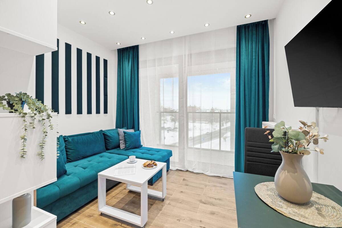 A modern living room with a teal sofa, white coffee table, and a large window with teal curtains.