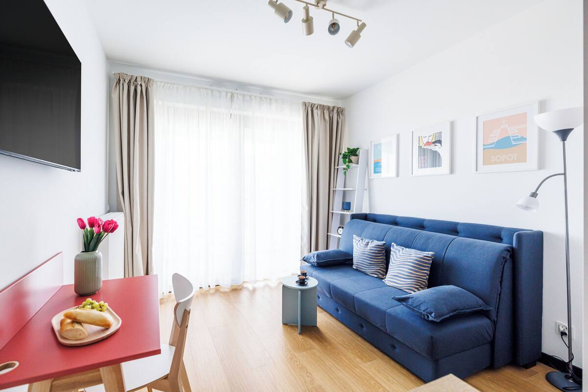 A modern living room with a blue sofa, a small round table with a vase of flowers, and a dining area with a red table and chairs.