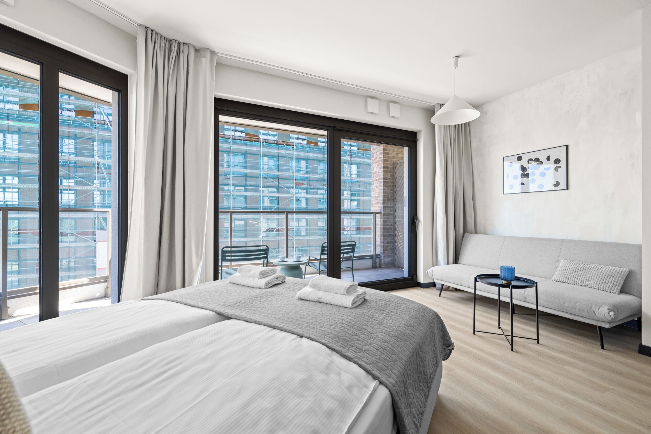 A modern hotel room with a large bed, a sofa, a small table, and a balcony with a view of a building under construction.