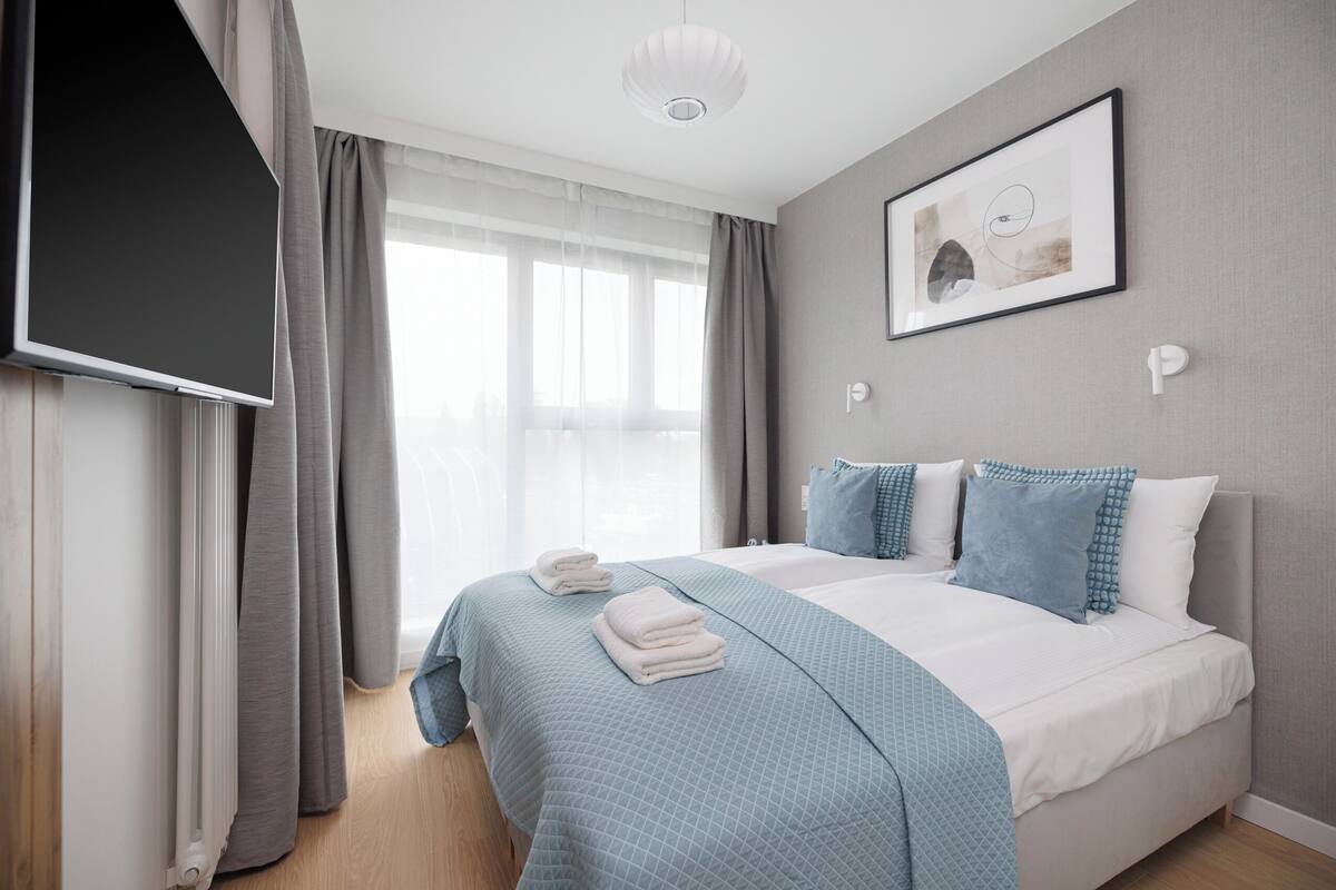 A modern hotel room with a large bed, a flat-screen TV, and a window with sheer curtains.