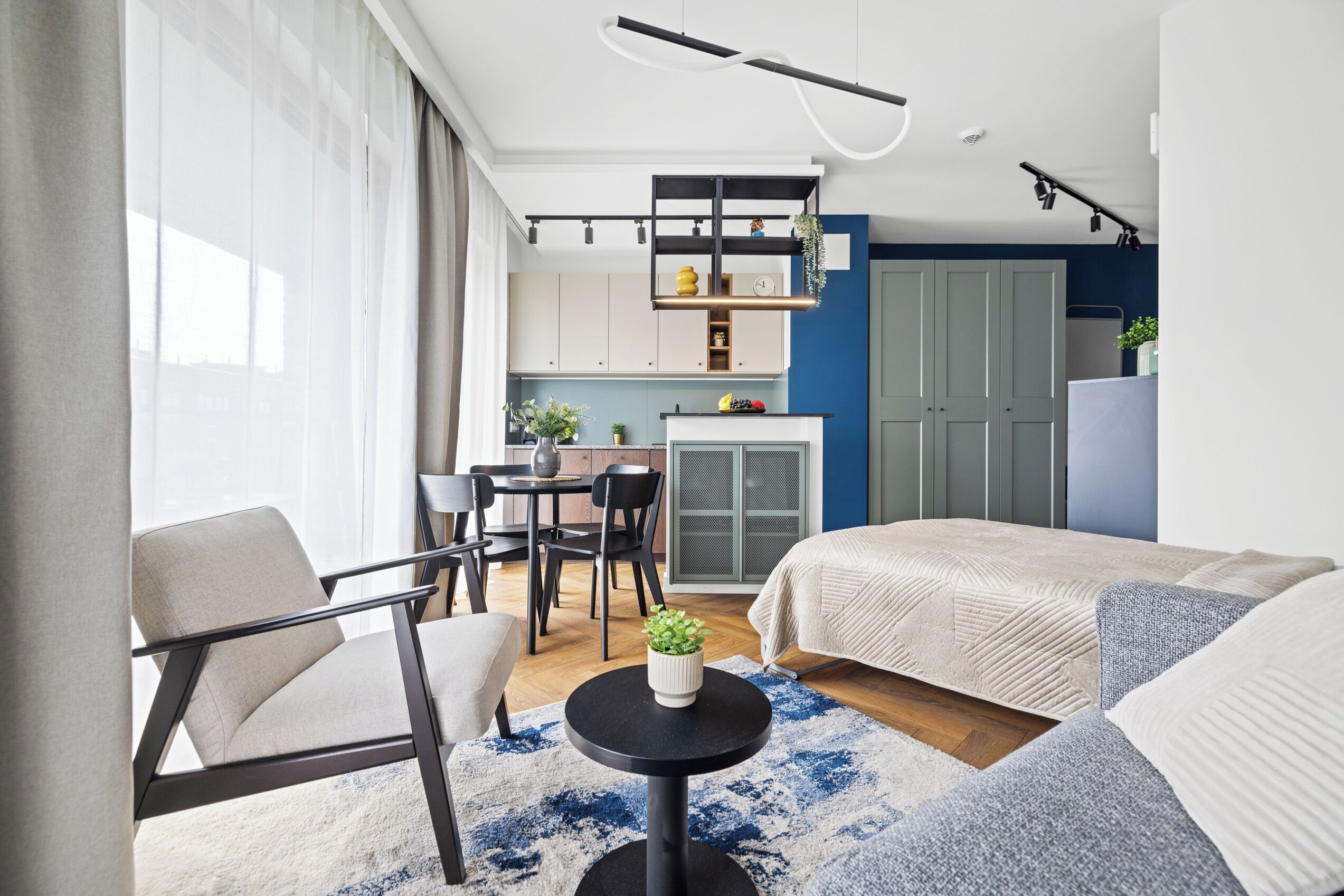 A modern hotel room with a bed, a dining table, chairs, a small round table with a plant, and a kitchenette.