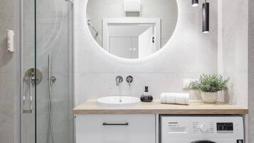Ett modernt badrum med en dusch med glasväggar, ett handfat med en spegel ovanför, en tvättmaskin och en krukväxt.