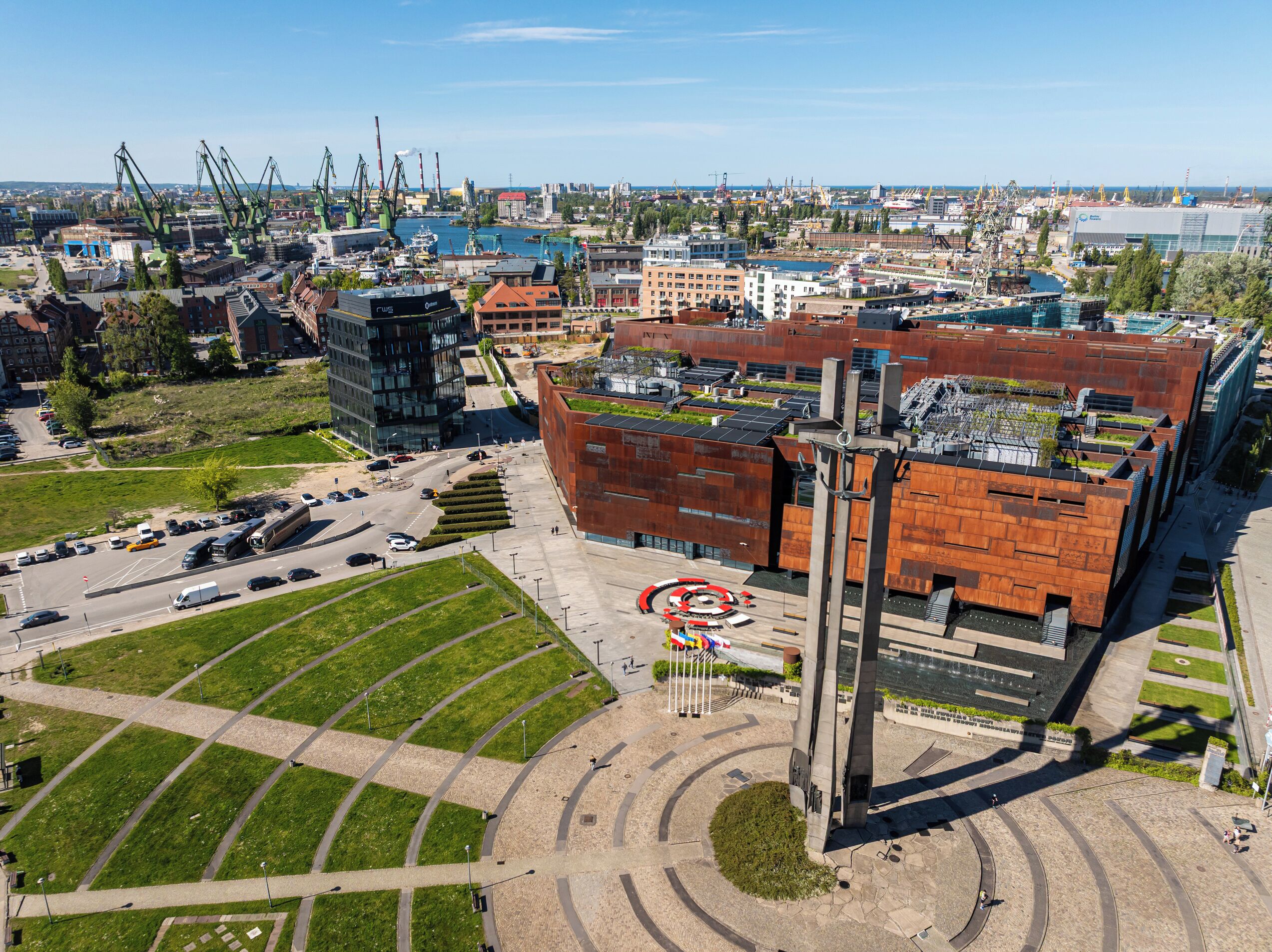 Vista aérea de una zona urbana moderna con un edificio destacado de diseño arquitectónico único, rodeado de espacios verdes y un puerto concurrido con grúas.