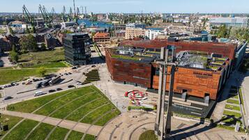 Vista aérea de una zona urbana moderna con un edificio destacado de diseño arquitectónico único, rodeado de espacios verdes y un puerto concurrido con grúas.