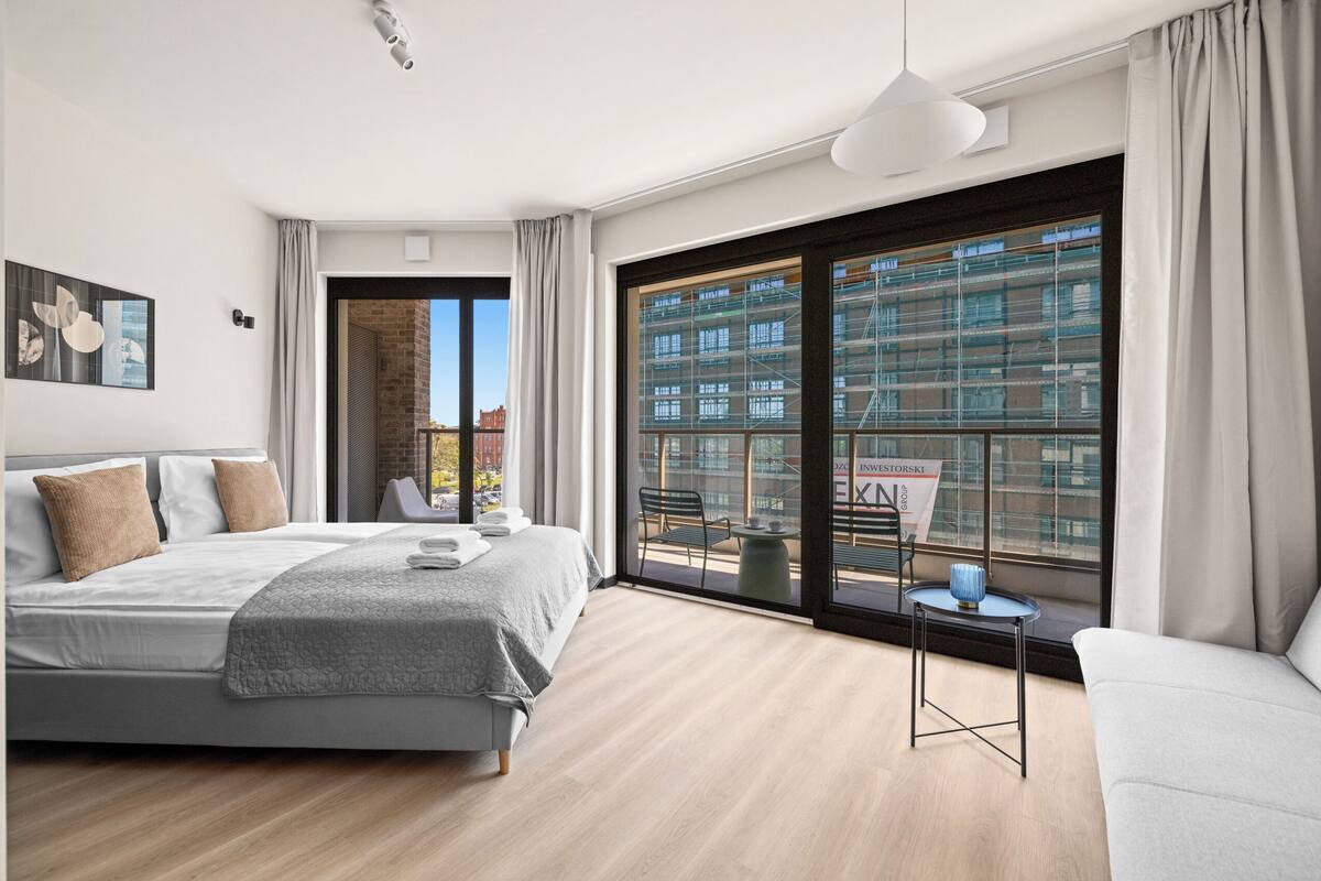 A modern bedroom with a large bed, a sofa, a small table, and a view of a cityscape through a large window.