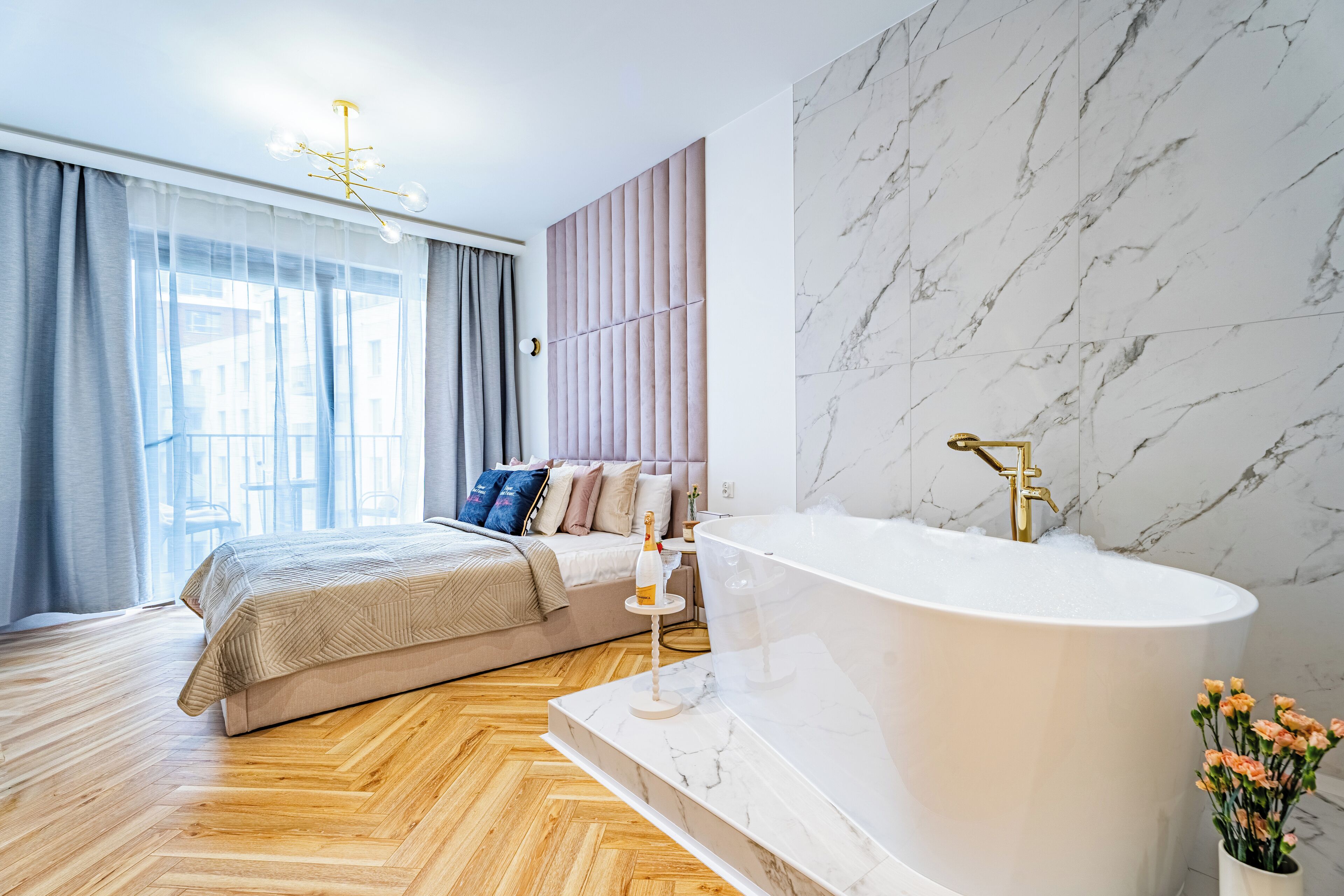 Une chambre moderne dotée d’un grand lit, d’une salle de bain en marbre avec une baignoire sur pieds et offrant une vue sur la ville grâce à de grandes fenêtres.