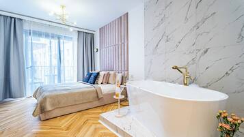 Une chambre moderne dotée d’un grand lit, d’une salle de bain en marbre avec une baignoire sur pieds et offrant une vue sur la ville grâce à de grandes fenêtres.