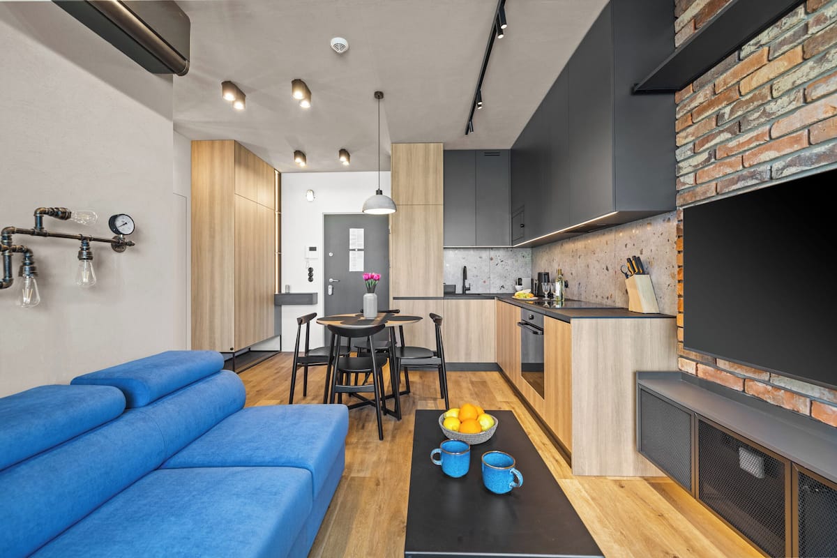 A modern kitchen with a brick accent wall, a dining area with a black table and chairs, and a living area with a blue sofa.
