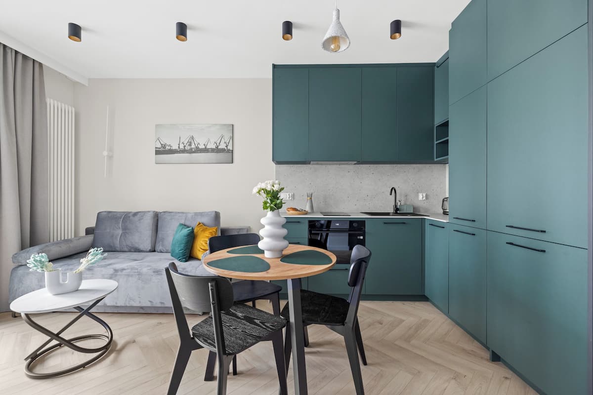 A modern kitchen with teal cabinets, a dining area with a round table and chairs, and a gray sofa with decorative pillows.