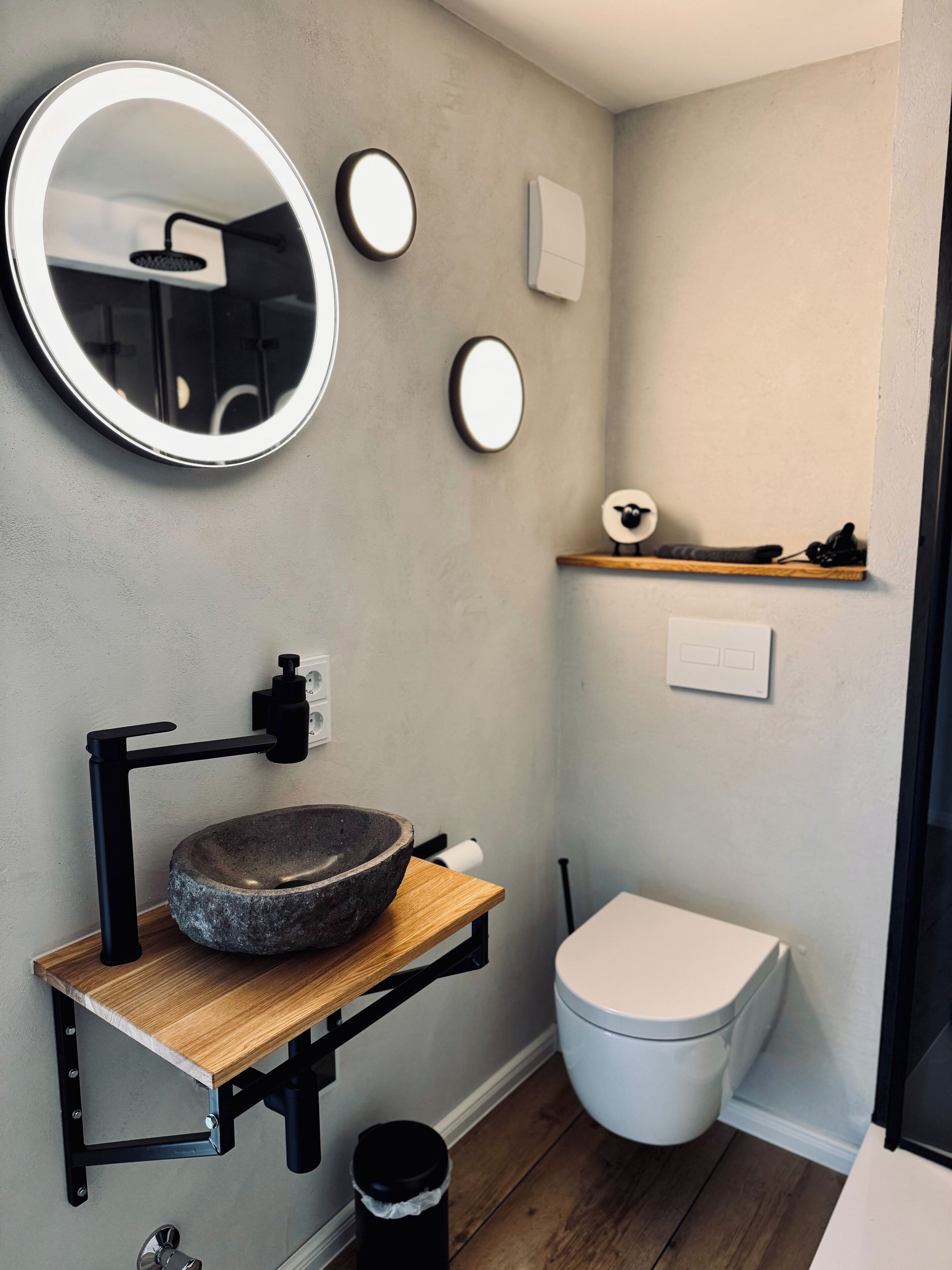 A bathroom with a round mirror, a stone sink, a toilet, and a shelf with a decorative item.