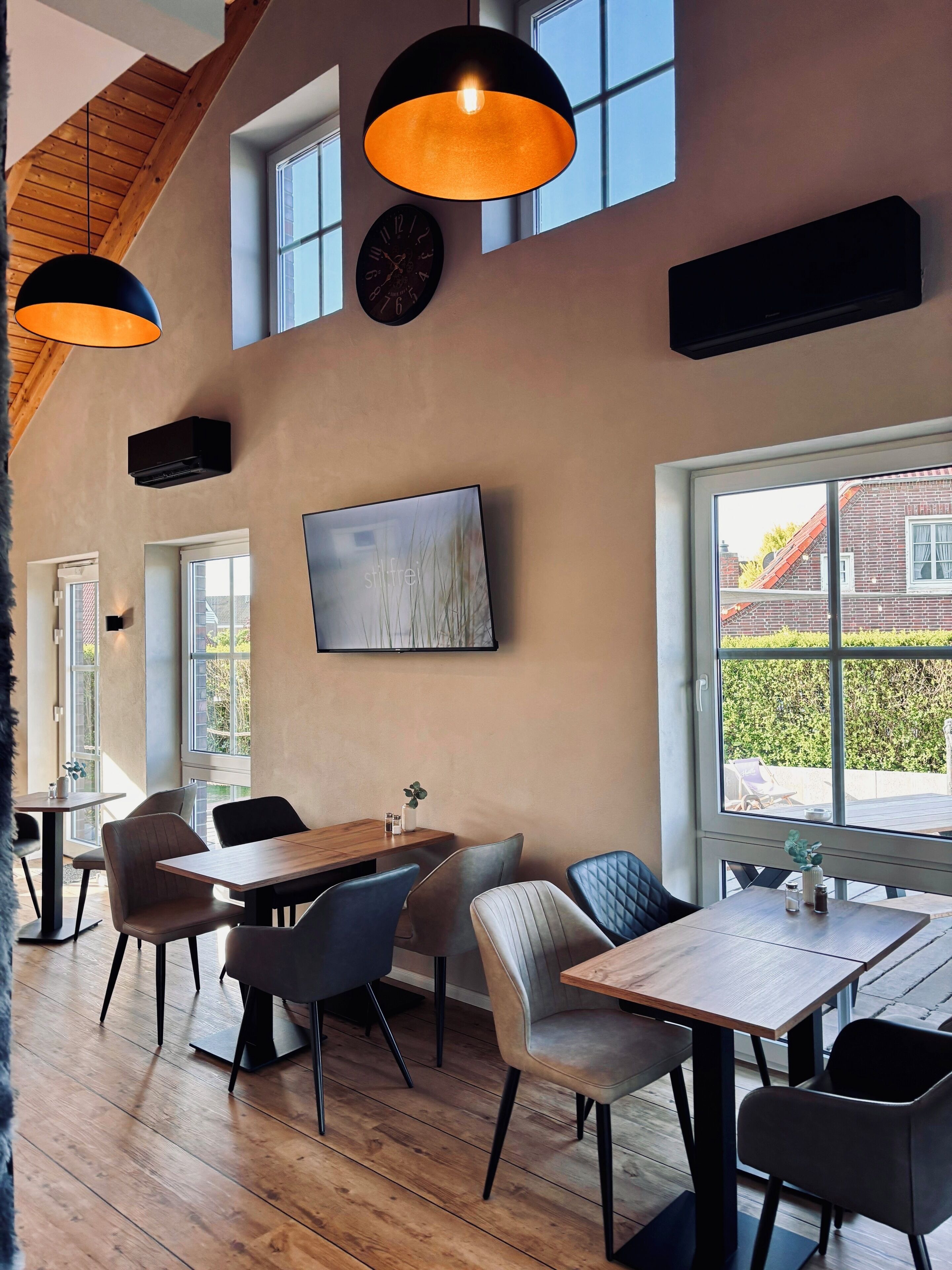 A modern dining area with wooden tables and chairs, pendant lighting, a large window with a view of a brick building, and a flat-screen TV mounted on the wall.