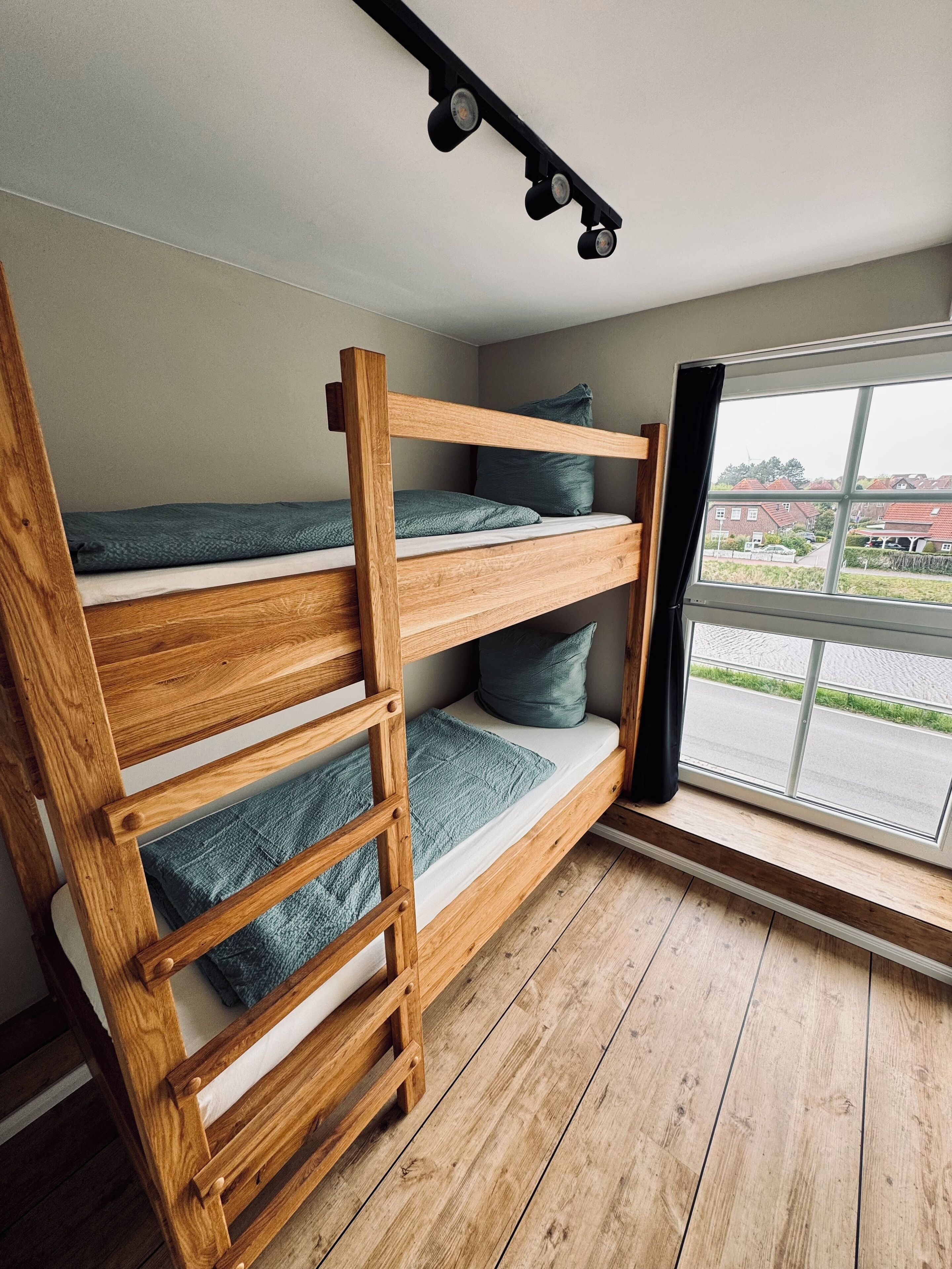 Una cama litera con escalera de madera, una ventana con vistas a casas y una lámpara en el techo.