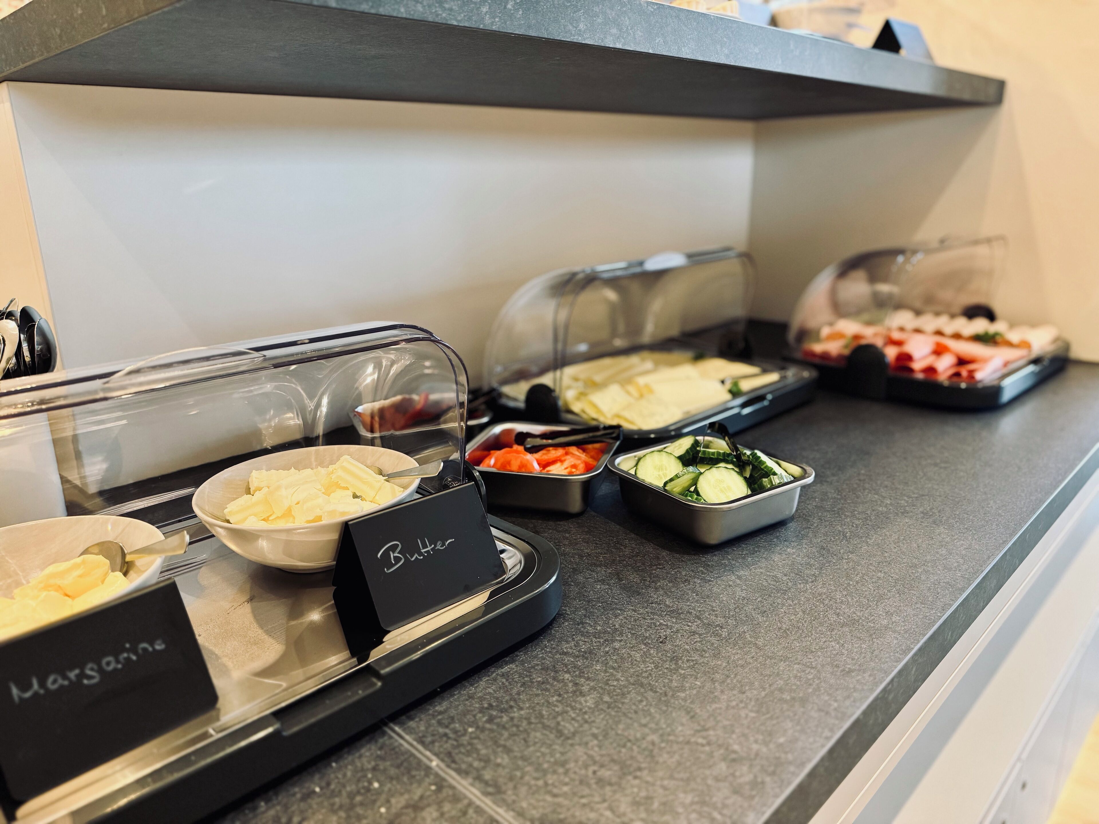 A buffet counter with various food items, including a bowl of cheese, a container of sliced cucumbers, and a tray of sliced meats.