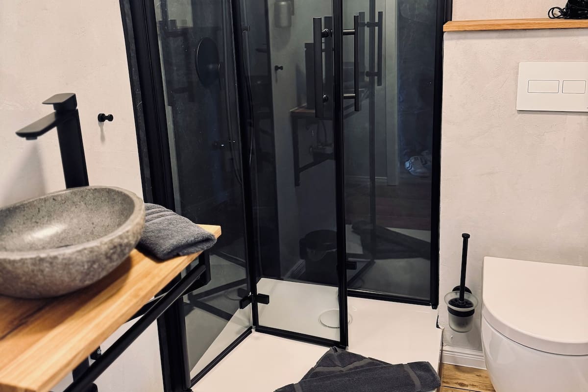 A modern bathroom with a glass-enclosed shower, a wooden vanity with a bowl sink, a toilet, and a small trash bin.