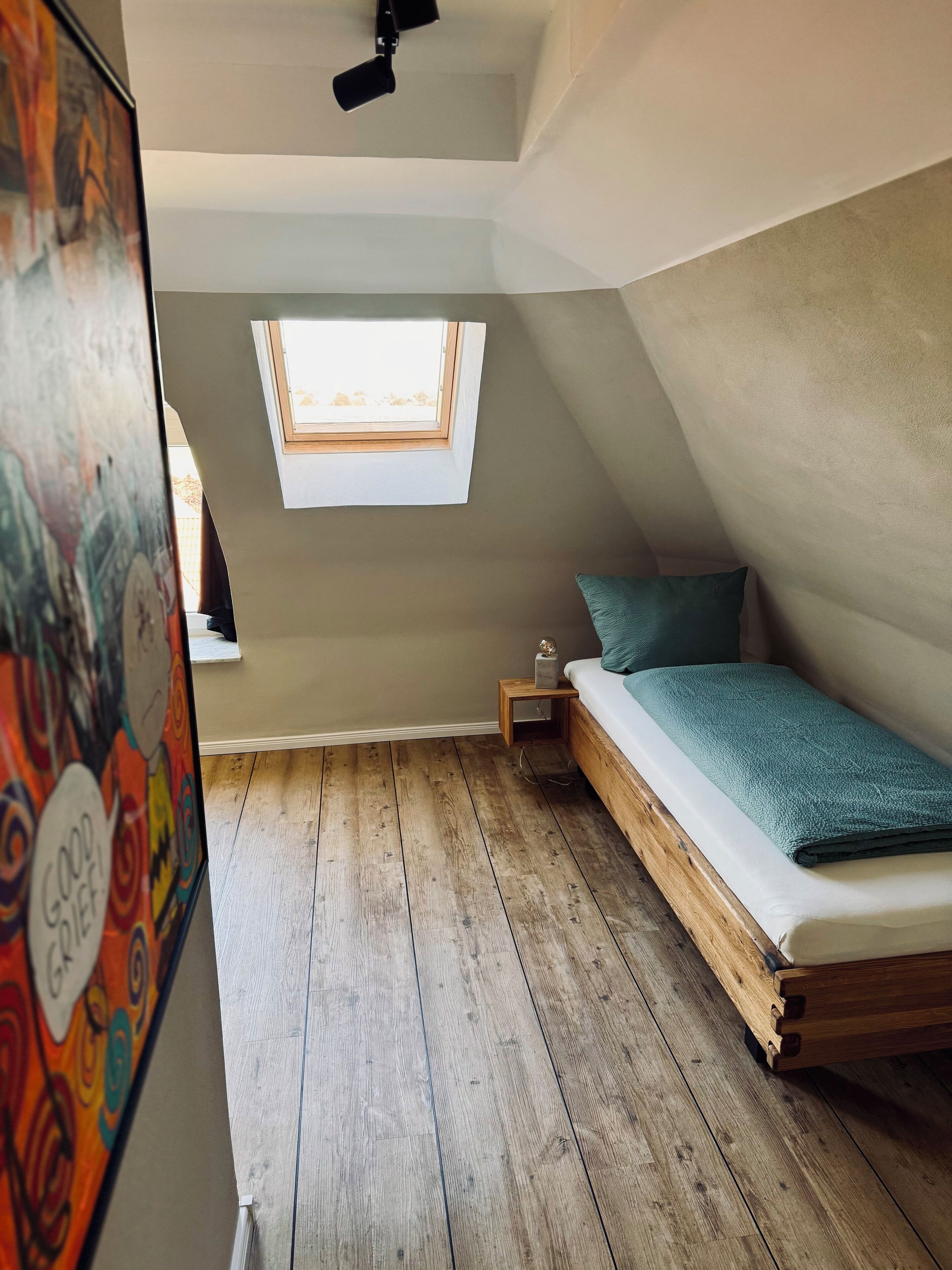 Um quarto pequeno com telhado inclinado, uma cama de solteiro, uma mesa de cabeceira, uma pequena mesa e uma janela com vista para o exterior.