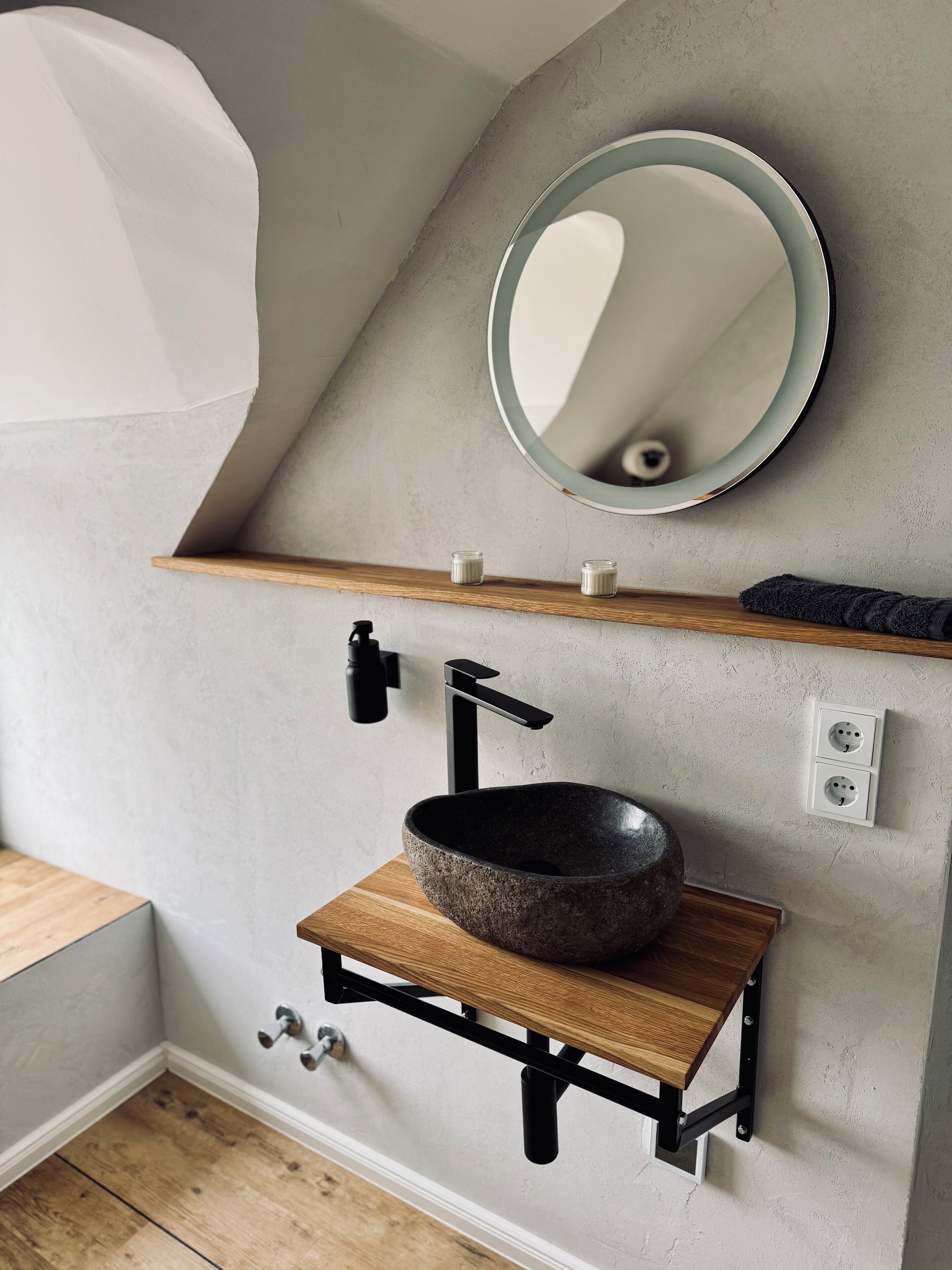 Ein Badezimmer mit einem Steinklopfbach, einem schwarzen Wasserhahn und einem Holzregal darüber.