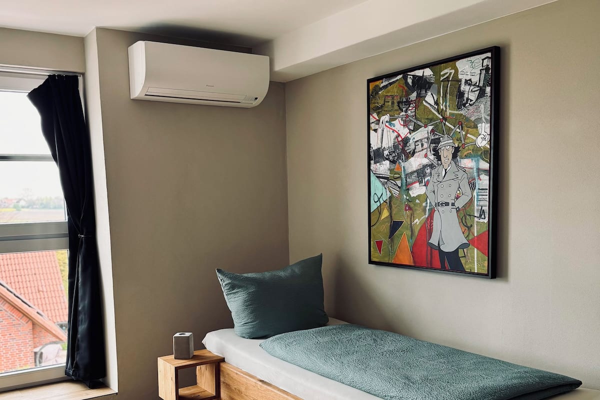 A compact room with a single bed, a small bedside table, a wall-mounted air conditioner, a window with curtains, and a colorful abstract painting.