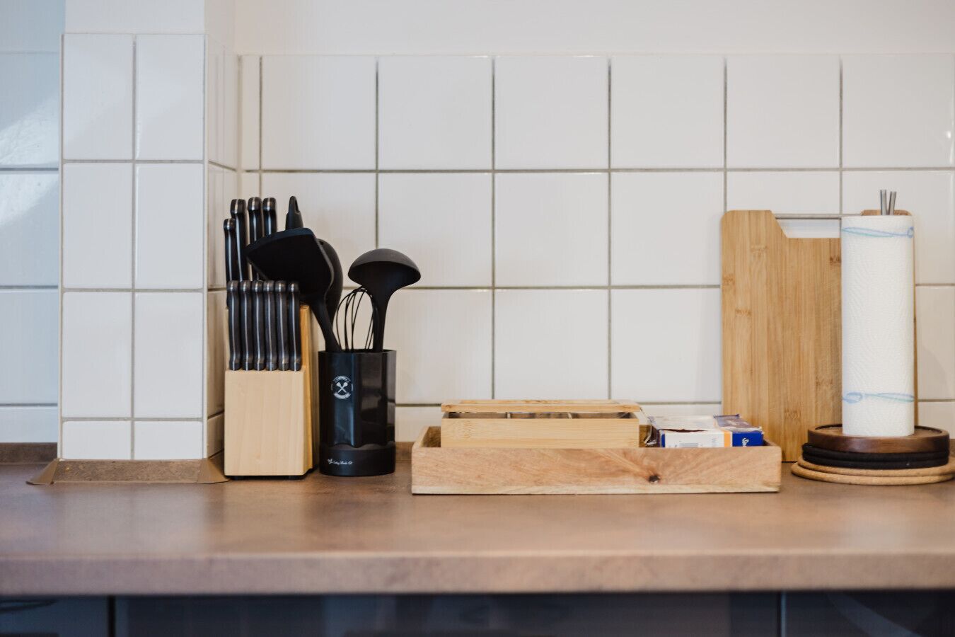 Een keukentrap met een messenblok, een zwarte container met keukengerei, een houten snijplank en een rol keukenpapier.