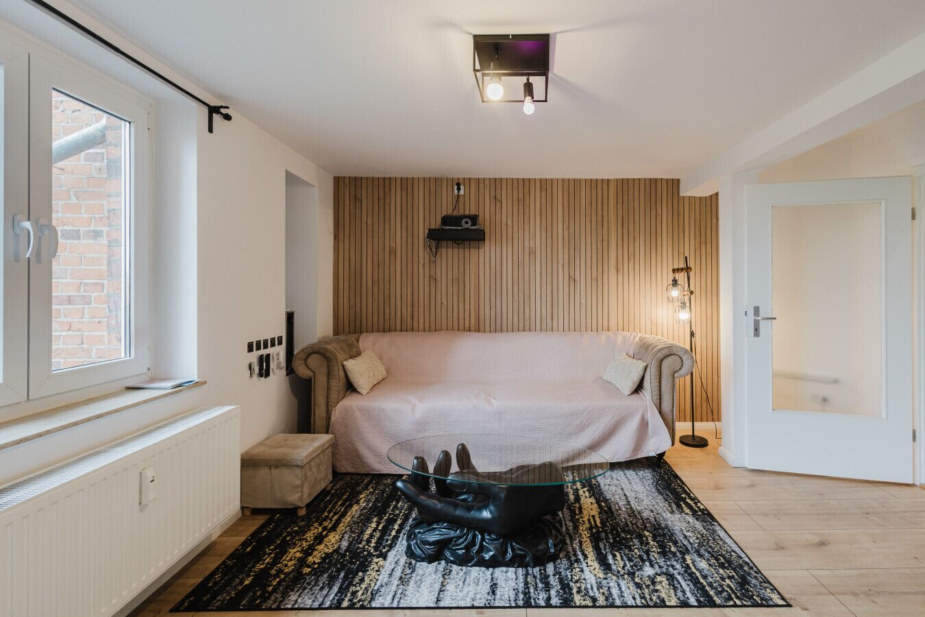 Quarto com cama, mesa de centro de vidro, radiador, janela e porta.