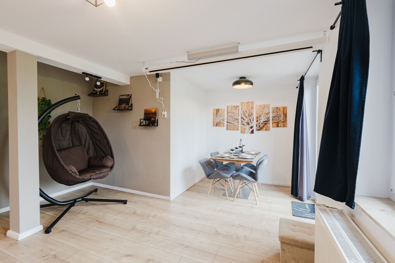 Un salon moderne avec un fauteuil suspendu, un coin repas avec une table et des chaises, et une grande fenêtre avec des rideaux.