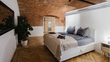 A bedroom with a herringbone wooden floor, a brick wall, and a bed with grey bedding and pillows.