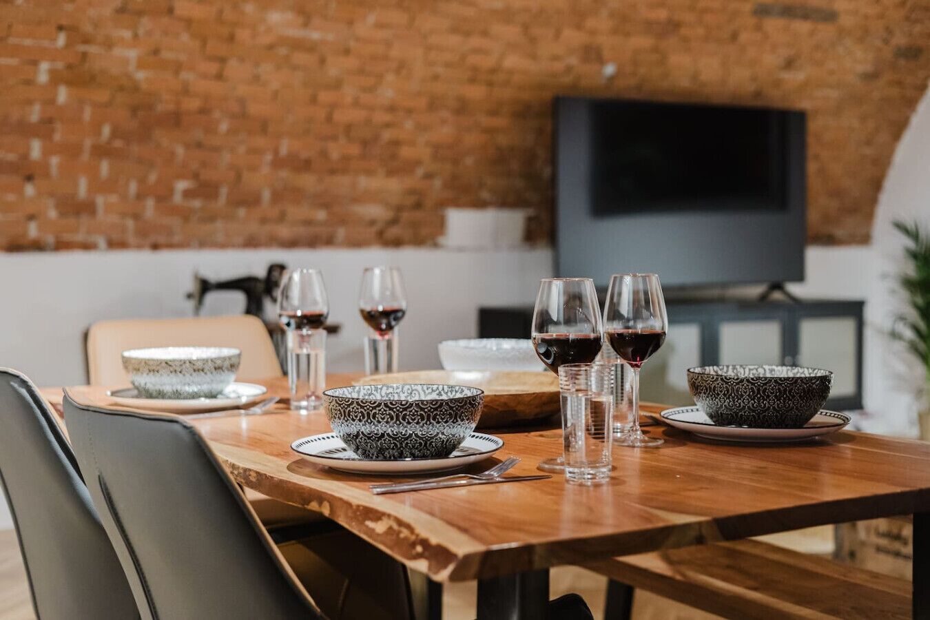 Una mesa de comedor dispuesta con fuentes y copas de vino, un televisor al fondo y una pared de ladrillo.