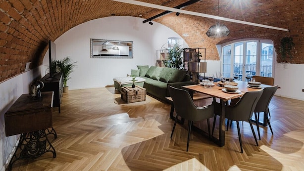 Spazioso loft con pareti in mattoni a vista, pavimento in legno a spina di pesce, zona pranzo con tavolo e sedie in legno, divano verde e un'ampia finestra che offre luce naturale.