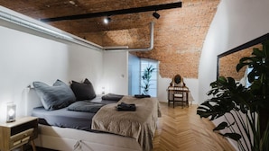 Camera da letto con pavimento in parquet a spina di pesce, letto con biancheria grigia, comodino in legno, specchio e una pianta in vaso.