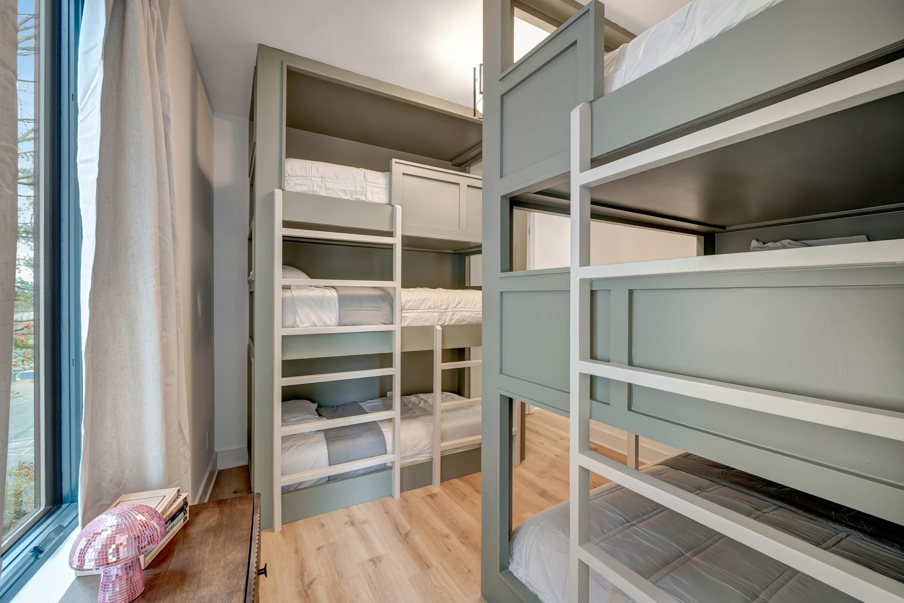 A room with bunk beds, a window with curtains, and a small table with a lamp.
