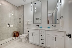 Un bagno moderno con box doccia in vetro, un water bianco, uno sgabello di legno, uno specchio grande e un mobile lavabo bianco con armadietti.
