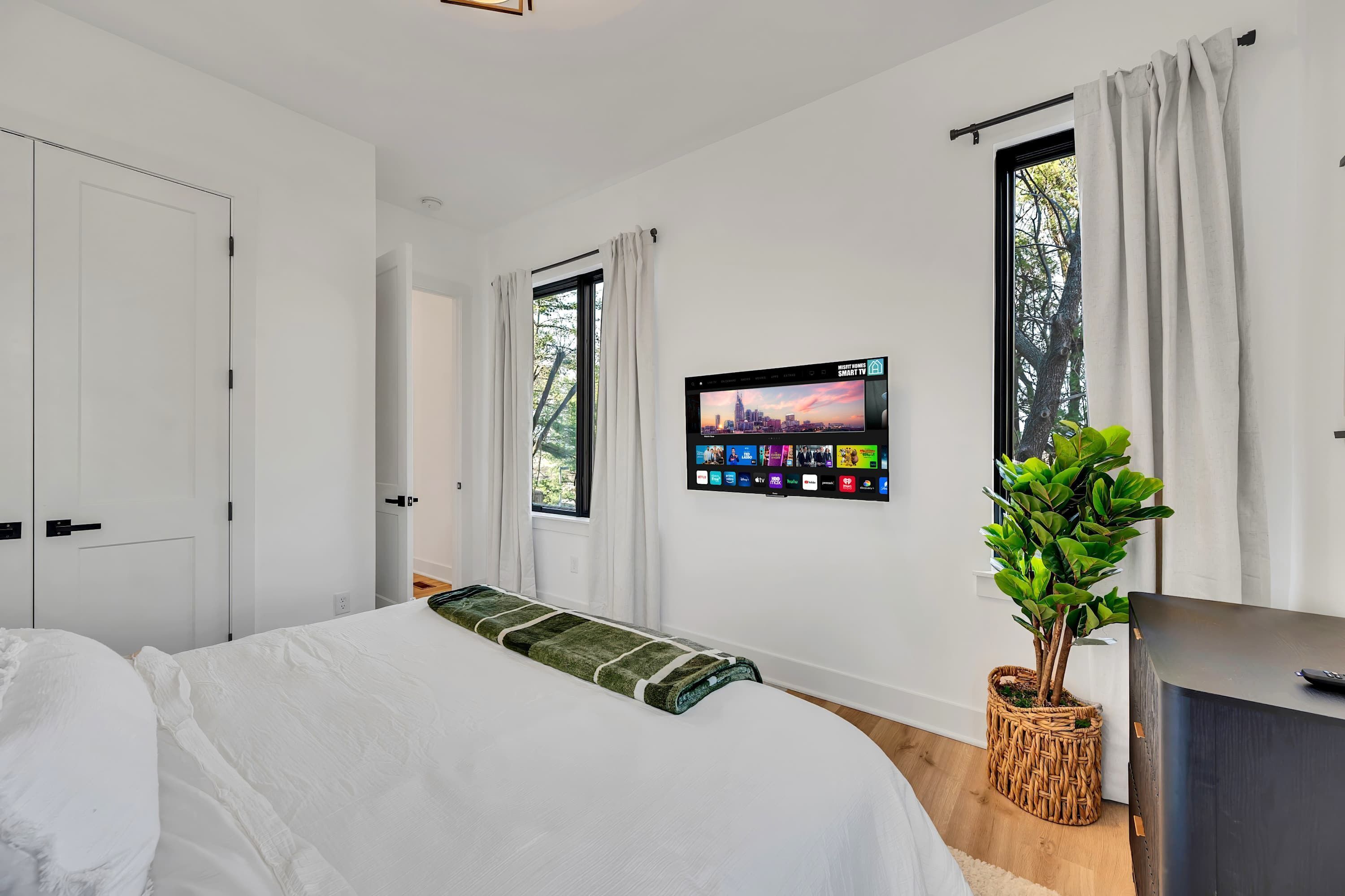 Un dormitorio moderno con una cama, un televisor montado que muestra un paisaje urbano y una planta en maceta.