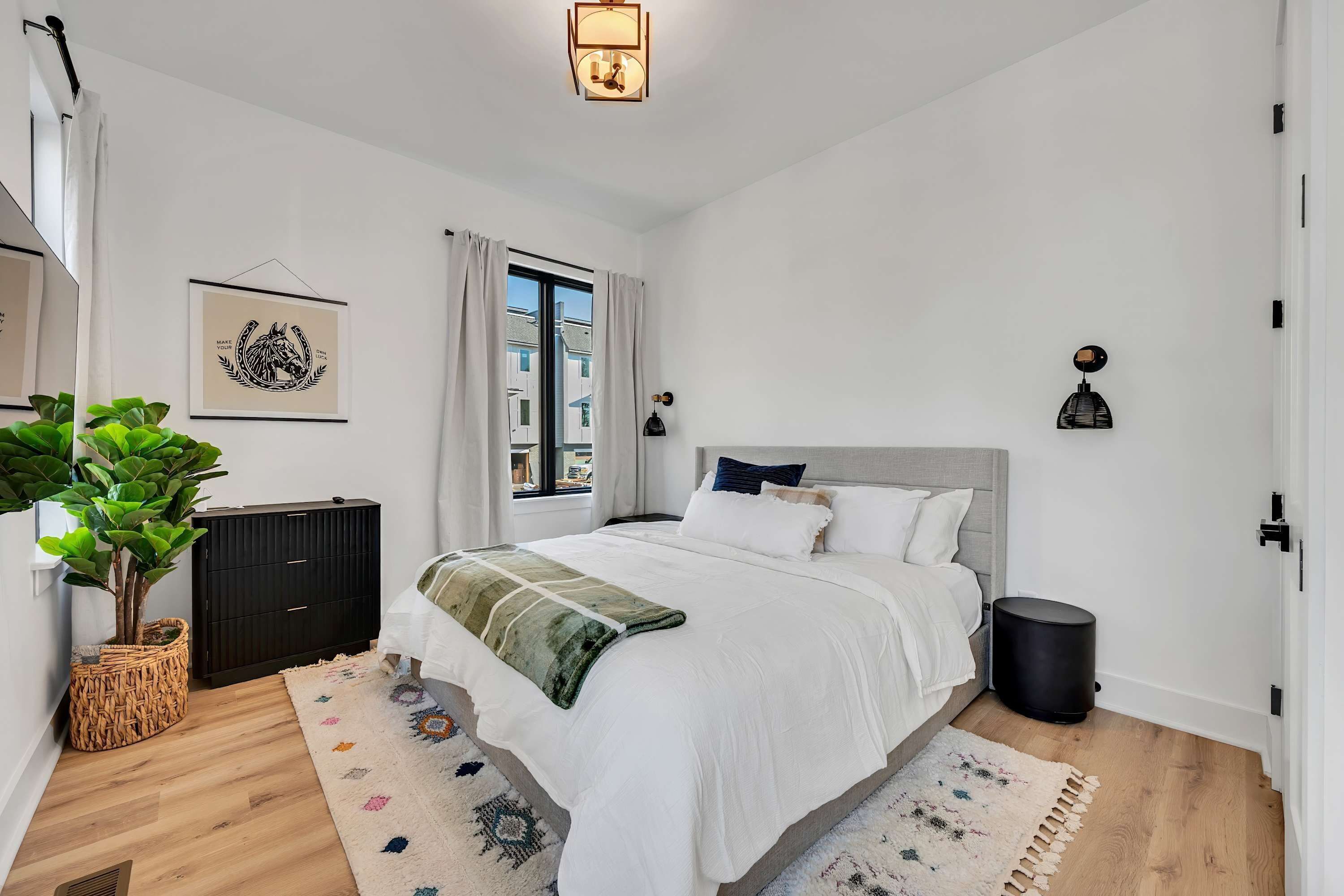 A bedroom with a bed, a potted plant, a black cabinet, a window, and a wall-mounted light fixture.