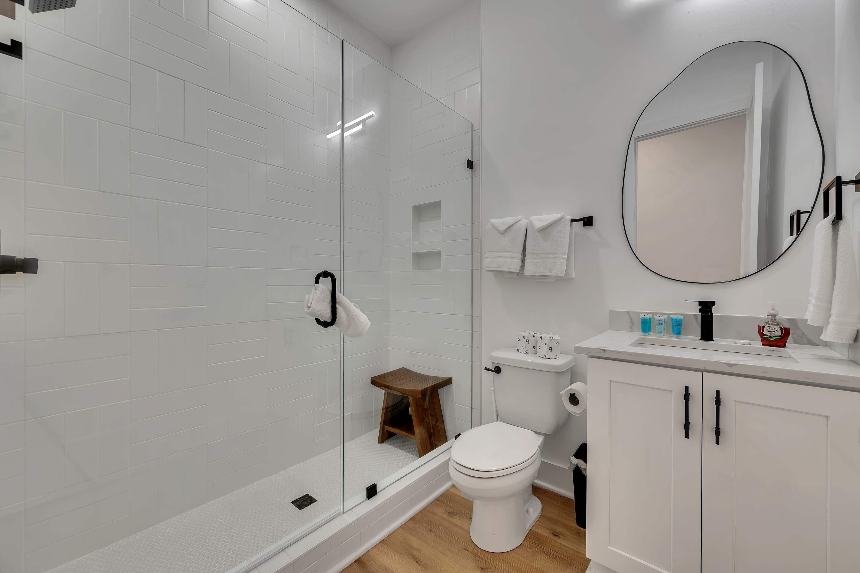 A modern bathroom with a glass shower enclosure, white tiles, a wooden stool, a white toilet, a vanity with a mirror, and a cabinet.