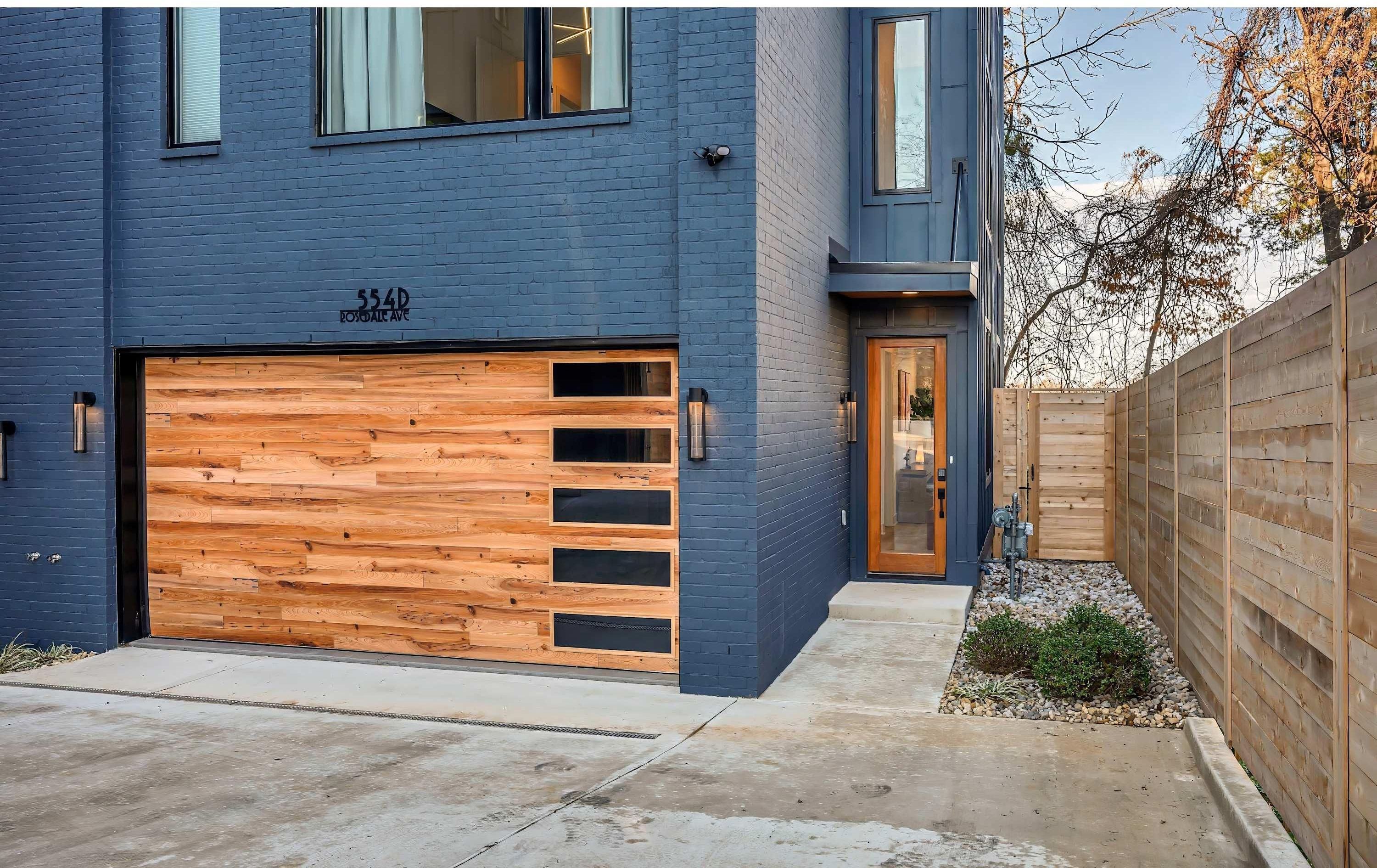 Une maison moderne à deux étages, avec une façade bleue, une porte de garage en bois et un petit jardin devant, délimité par une clôture en bois.