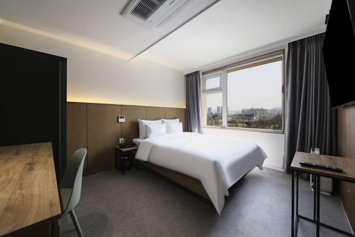 Habitación de hotel con cama, escritorio, silla, ventana y vista a la ciudad, y un televisor.