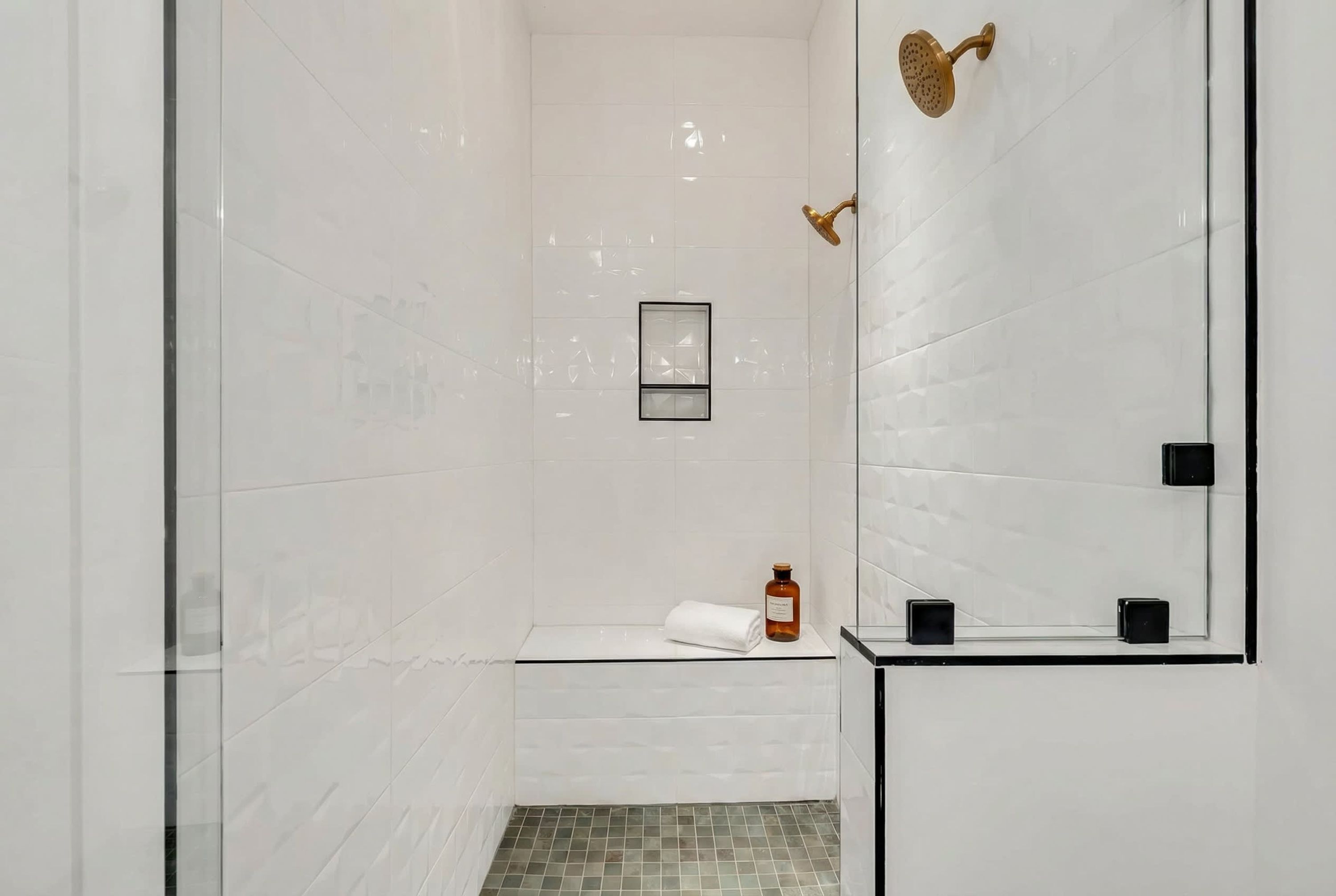 A modern bathroom with white tiled walls, a glass shower enclosure, a bench with a towel, and a framed picture on the wall.