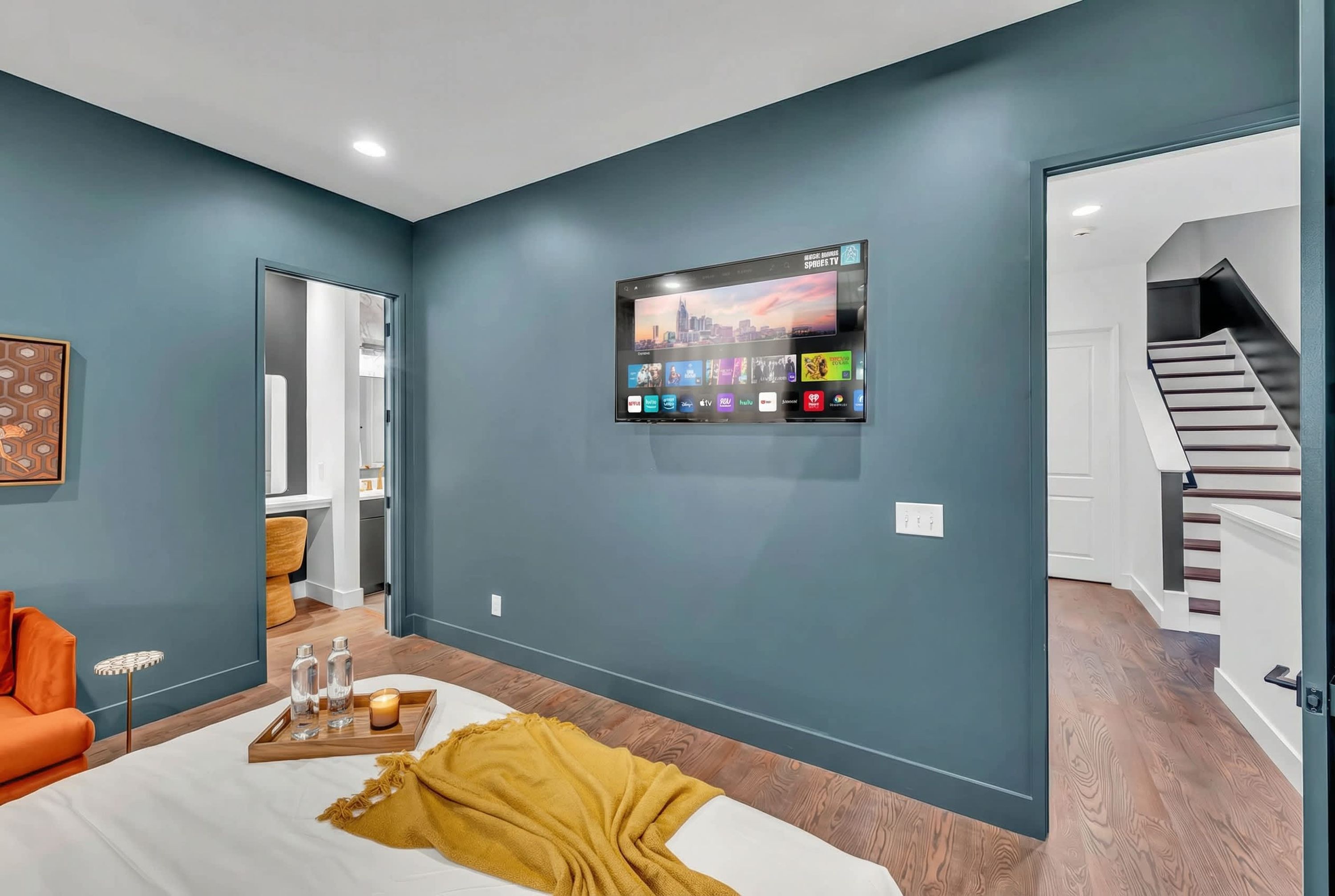 A modern hotel room with a large bed, a mounted TV displaying a cityscape, a wooden floor, and a staircase leading to another level.