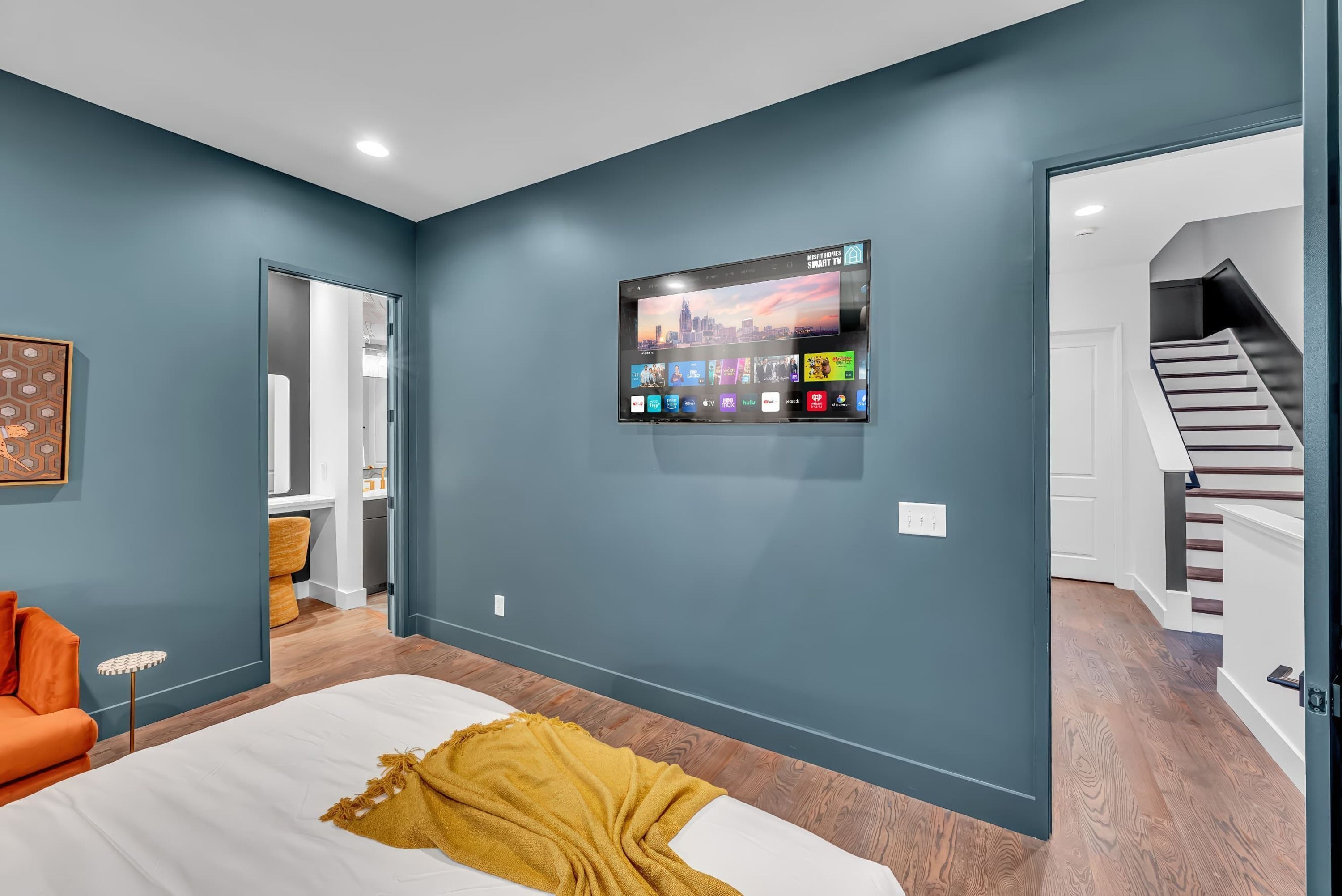 Ein modernes Schlafzimmer mit einem großen Bett, einem an der Wand montierten Fernseher, der verschiedene Sender zeigt, und einer Treppe, die zu einer anderen Ebene führt.