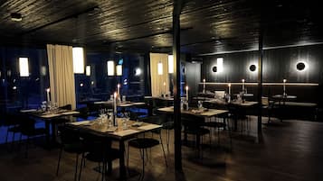 A dimly lit restaurant with tables set for dining, featuring candle-lit centerpieces and a view of the night sky through large windows.