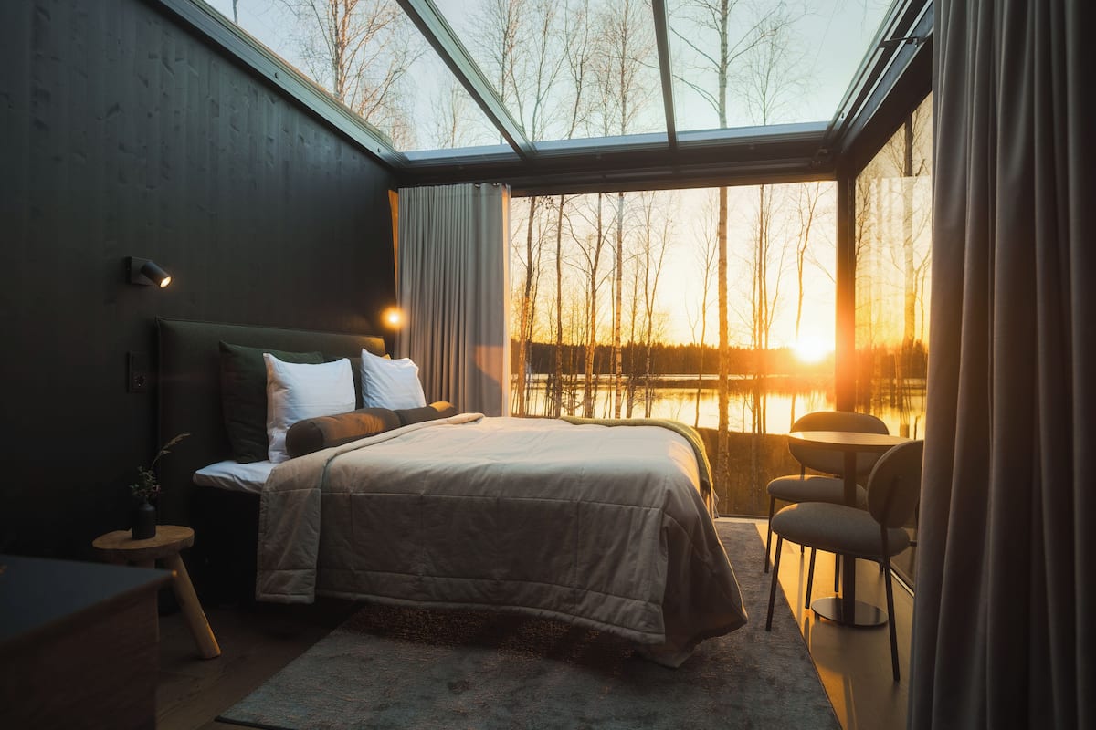 Quarto com uma cama grande, mesa de cabeceira, cadeira e vista para um lago ao pôr do sol.