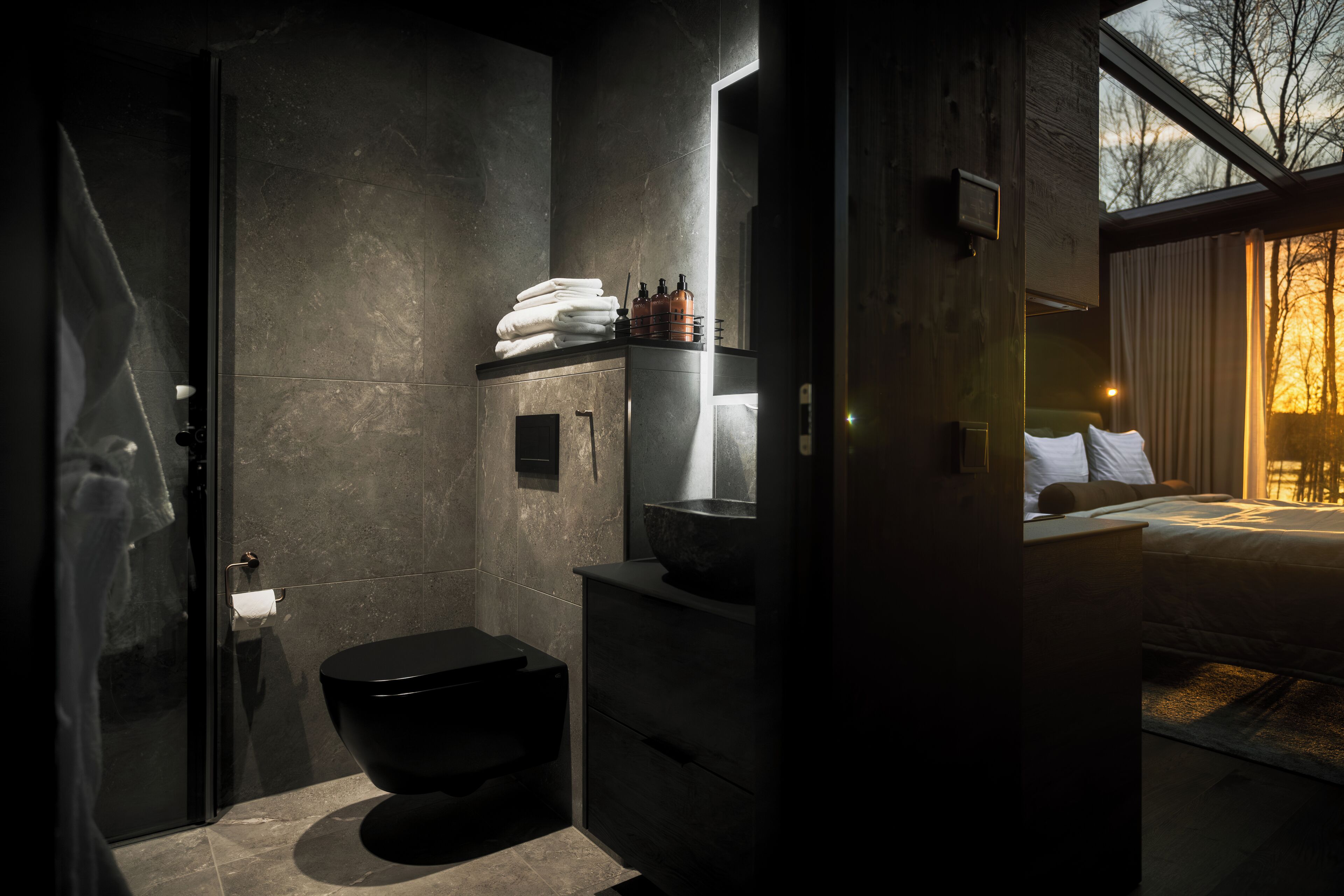 A modern bathroom with a black toilet, a sink, and a mirror. There is a towel rack with folded towels and a bottle of hand soap.