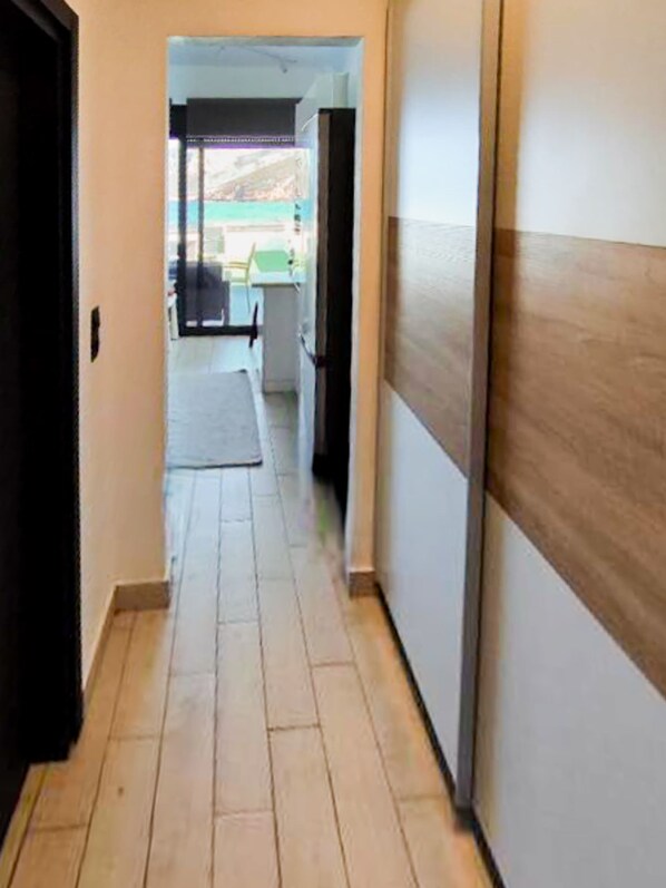 Un couloir doté d’un plancher en bois et de murs blancs, qui mène à une pièce avec une fenêtre donnant sur un bâtiment à l’extérieur.